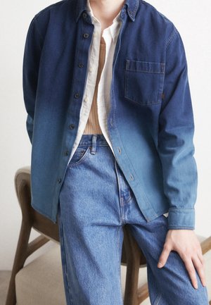 Osoba ubrana w niebieską koszulę z denimu z efektem ombre, na białej koszuli zapinanej na guziki i jasnobrązowym topie, w połączeniu z wysokim stanem niebieskich dżinsów, siedząca na krześle.