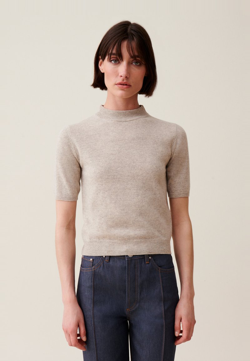 Maglione beige a maniche corte con collo alto; texture lavorata a maglia; design attillato; indossato con jeans in denim scuro con cuciture più chiare.