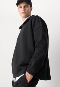 Felpa nera con collo alto, chiusura con zip, vestibilità ampia, polsini a costine, texture spazzolata, indossata sopra una camicia bianca.