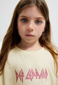 Gelbes Baumwoll-T-Shirt mit dem Schriftzug "DEF LEPPARD" in Rot, getragen von einem Kind mit langen Haaren und neutralem Gesichtsausdruck.