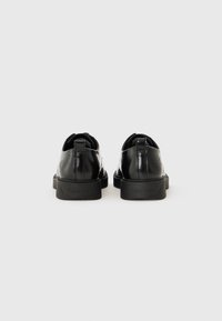Chaussures en cuir noir avec une finition brillante, semelle en caoutchouc texturée et logo en relief au niveau du talon. Bout arrondi et tirant à l'arrière pour un enfilage facile.