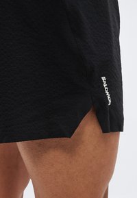 Κοντινό πλάνο στο πάνω μέρος μηρού ενός ατόμου που φοράει μαύρο αθλητικό σορτς με υφή, με μια μικρή πλαϊνή σχισμή και το λογότυπο "Salomon" σε λευκό.