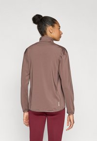 Barna, hosszú ujjú sportdzseki nedvszívó anyagból, magas nyakkal és rugalmas mandzsettával, bordó leggingsszel párosítva.