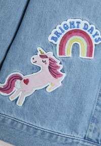 Denim azul claro con parches bordados: un unicornio rosa y un arcoíris con el texto "DÍAS BRILLANTES," presentando varios colores pastel.
