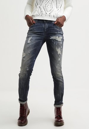 Personne portant un jean skinny foncé usé, un pull en tricot blanc et des bottes bordeaux à lacets, debout les mains dans les poches.