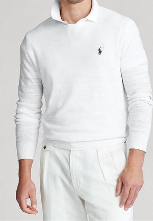 Homme portant un polo en maille blanche à manches longues avec un petit logo bleu marine sur la poitrine et un pantalon blanc plissé.