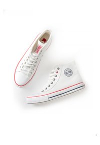 White high-top canvas sneakers with red accents, featuring a round logo patch, contrasting blue stripes on the sole, and metal eyelets.