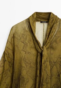Blouse vert olive avec motif imprimé serpent et un nœud lâche au col, réalisée en tissu léger.