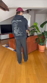 Melnā kapuce ar ziedu rakstu aizmugurē, apvienota ar brīviem pelēkiem biksēm. Siltas koka grīdas un liela zaļa augstsiena redzama.