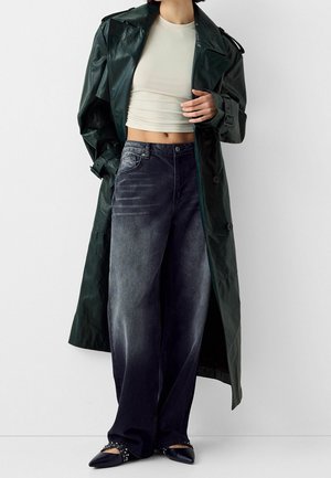 Personne portant un long trench-coat en cuir vert foncé, un top court crème, un jean ample noir délavé et des ballerines noires pointues cloutées.