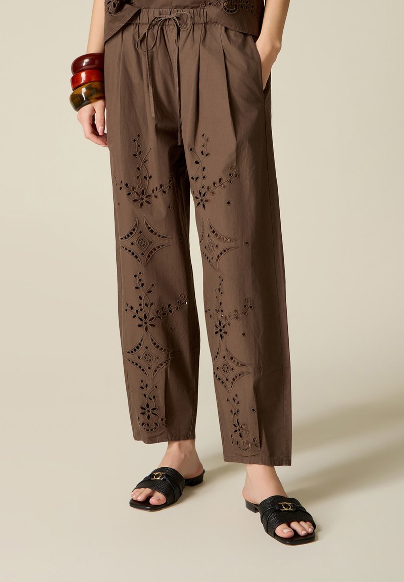 Pantalones marrones de pierna ancha con bordado floral calado, combinados con sandalias negras tipo slide y pulseras gruesas apiladas en una muñeca.