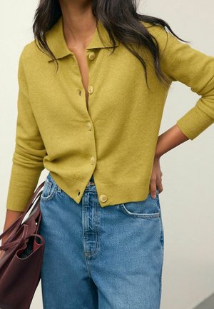 Femme portant un cardigan jaune moutarde boutonné et un jean bleu taille haute, tenant un sac à main en cuir marron avec la main sur la hanche.