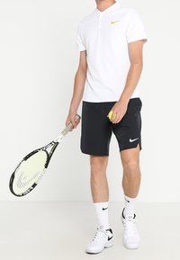 Camisa polo blanca con el logo dorado de Nike, combinada con pantalones cortos negros. Calcetines blancos y zapatillas, sosteniendo una raqueta de tenis y una pelota de tenis amarilla.