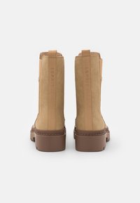 Bottines en daim beige avec des panneaux élastiques sur les côtés, semelles plates en caoutchouc marron et un subtil logo embossé à l'arrière.