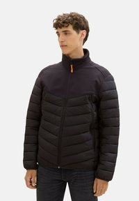 Czarna kurtka puchowa z pikowanym wzorem, wysokim kołnierzem i dwiema kieszeniami na zamek. Zamek ma pomarańczowy uchwyt. Nosi się ją na ciemnej koszuli.