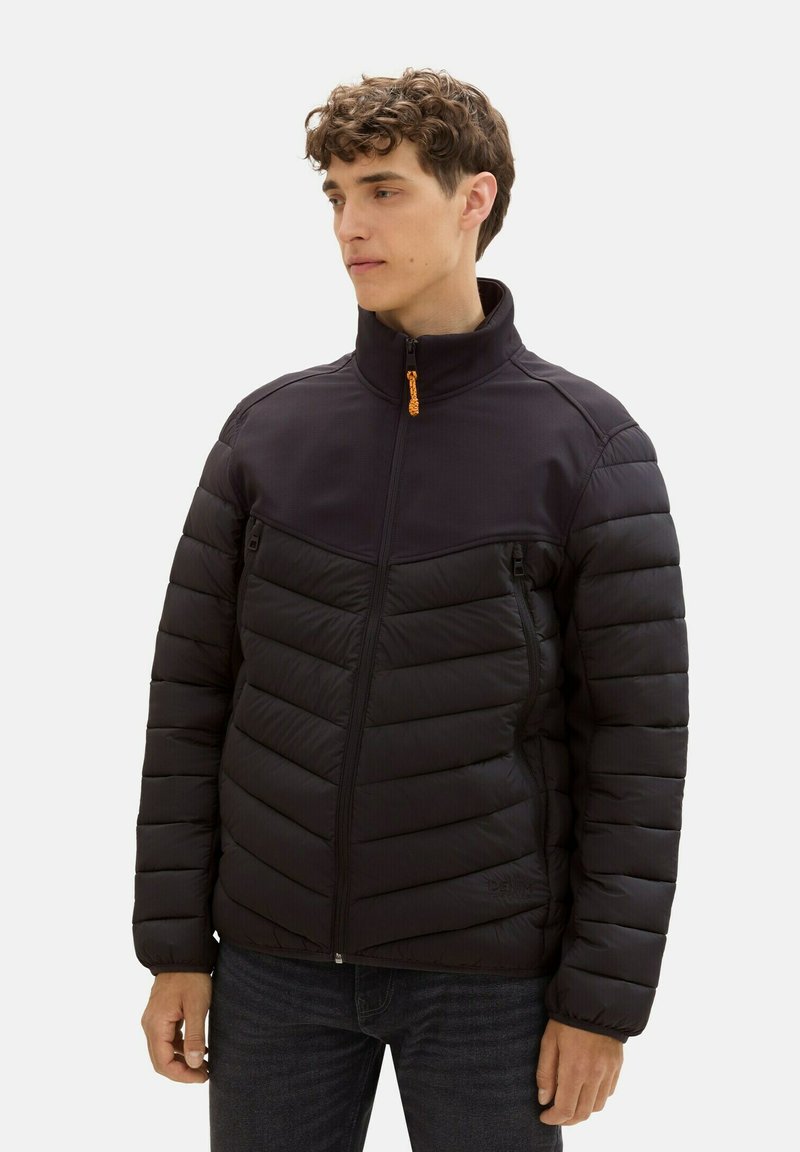 Czarna kurtka puchowa z pikowanym wzorem, wysokim kołnierzem i dwiema kieszeniami na zamek. Zamek ma pomarańczowy uchwyt. Nosi się ją na ciemnej koszuli.