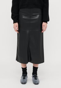 Saia midi de couro preta com uma silhueta ajustada, textura suave e um detalhe de costura a correr verticalmente pelo centro. Acentuada com sapatos cinzentos.