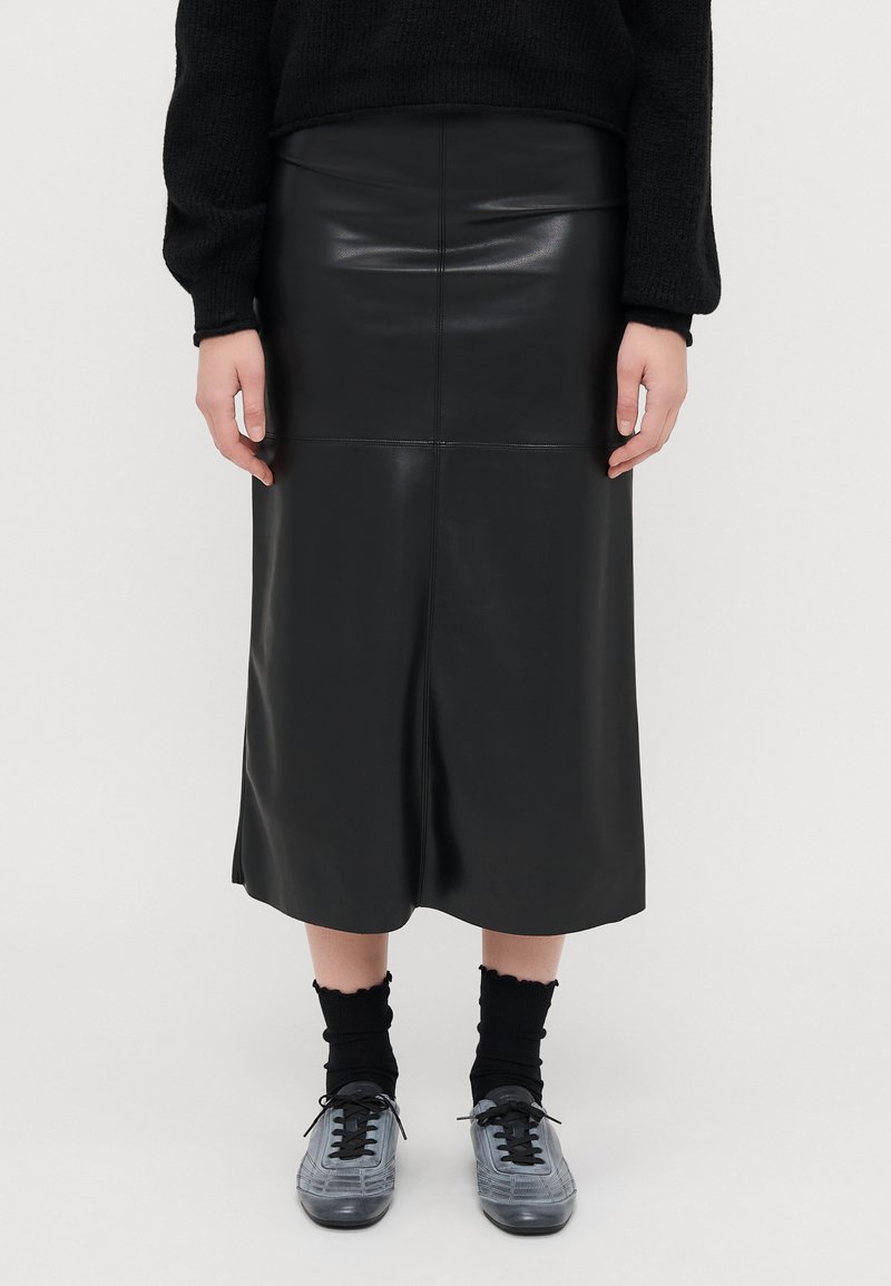 Saia midi de couro preta com uma silhueta ajustada, textura suave e um detalhe de costura a correr verticalmente pelo centro. Acentuada com sapatos cinzentos.