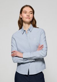 Camicia button-up azzurro chiaro con un leggero motivo a quadretti, colletto a punta e maniche lunghe con polsini abbottonati. Dettaglio del logo discreto sul petto.