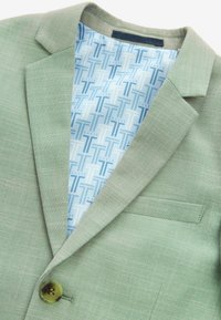 Blazer verde chiaro in tessuto texturizzato, chiusura con un bottone singolo e rivestimento interno blu a motivo con loghi. Include un dettaglio sulla tasca del petto.