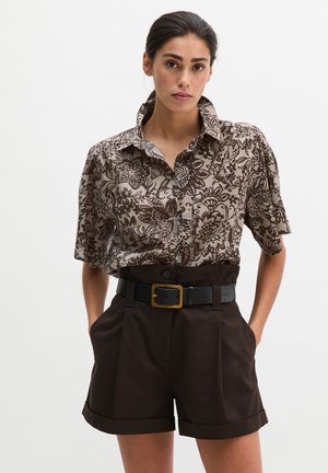 Woman wearing a short-sleeve floral print blouse tucked into high-waisted dark shorts with a black belt, standing with hands in pockets.
