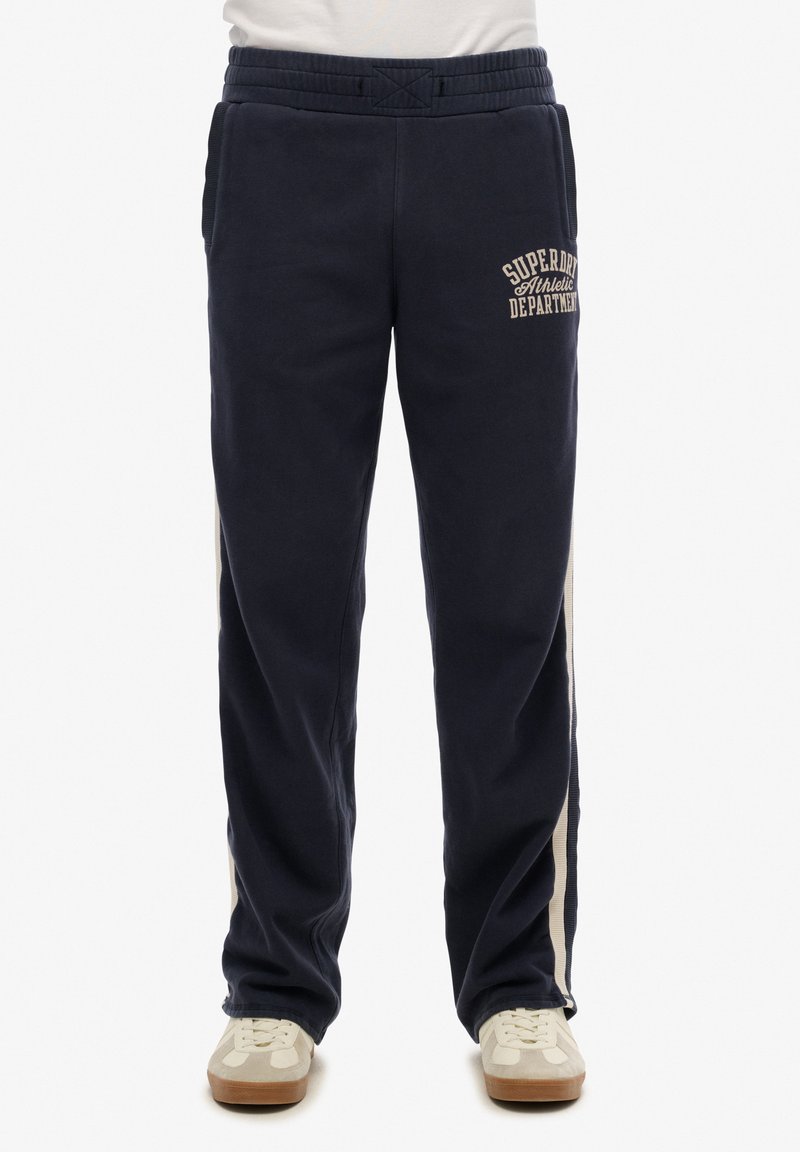 Pantalones de chándal azul marino con cintura elástica, bolsillos laterales y detalles de franjas en un tono beige claro. Presenta texto con el logo en el muslo superior. Textura suave.