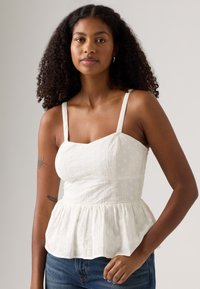 Top peplum blanc avec bretelles réglables, présentant des motifs floraux brodés et un corsage ajusté, associé à un jean en denim bleu.