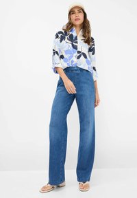 Light blue and navy floral patterned oversized shirt, paired with wide-leg blue denim jeans and beige flip-flops. White backdrop.