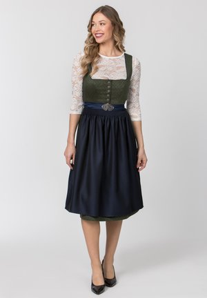 Frau lächelnd, trägt traditionelles grünes und dunkelblaues Dirndl mit weißen Spitzenärmeln und schwarzen Absätzen, steht vor einem einfarbigen Hintergrund.