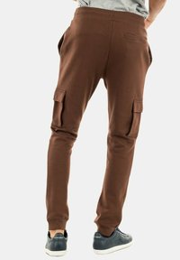 Pantalones deportivos marrones con una cintura acanalada y bolsillos laterales tipo cargo. Presentan piernas ajustadas y costuras en contraste, combinados con zapatillas oscuras.