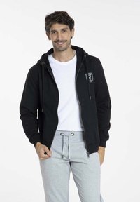 Haut à capuche zippé noir avec une poche avant et un cordon de serrage, porté par-dessus un t-shirt blanc. Un pantalon de survêtement gris complète la tenue.