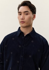 Marineblå kordfløyelskjorte med små broderte sirkulære mønstre, knappelukking foran, og krage. Har en diskret logo på brystet.