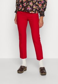 Pantalons en coton rouge à coupe slim et longueur cheville, associés à des sabots marron avec des clous dorés et des chaussettes blanches montantes.