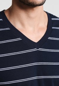 Homme portant un t-shirt V en bleu marine avec de fines rayures horizontales blanches, visibles du cou à la partie supérieure de la poitrine.