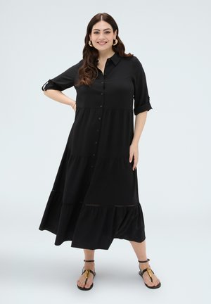 Donna sorridente che indossa un vestito midi nero abbottonato con maniche arrotolate e sandali neri con decorazioni dorate, in piedi su uno sfondo chiaro uniforme.