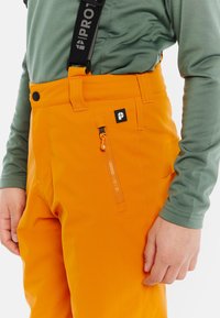 Heldere oranje sneeuwbroeken met verstelbare banden, een ritszak en een gladde textuur. Gecombineerd met een groene longsleeve shirt.