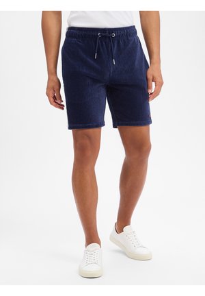 Mann trägt marineblaue Cord-Shorts mit Kordelzug und weiße Sneakers, steht vor einem schlichten weißen Hintergrund.