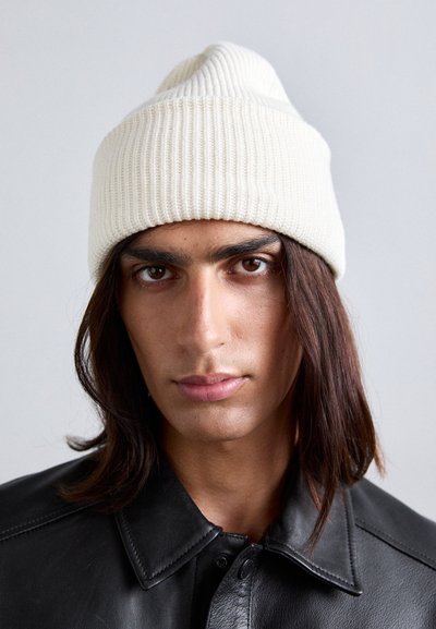 Person with long dark hair wearing a ribbed white knit beanie and a black leather jacket facing forward against a plain background.