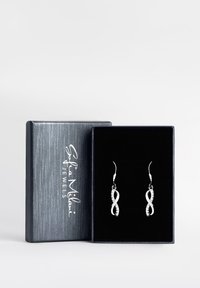 Silver infinity-formade örhängen med glänsande utsmyckningar, paketerade i en mörkblå texturerad låda med varumärket visat.