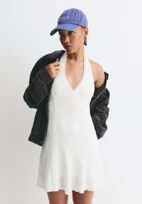 Robe halter blanche texturée avec dentelle, associée à une veste en velours côtelé noire et une casquette de baseball bleue ornée de texte brodé.