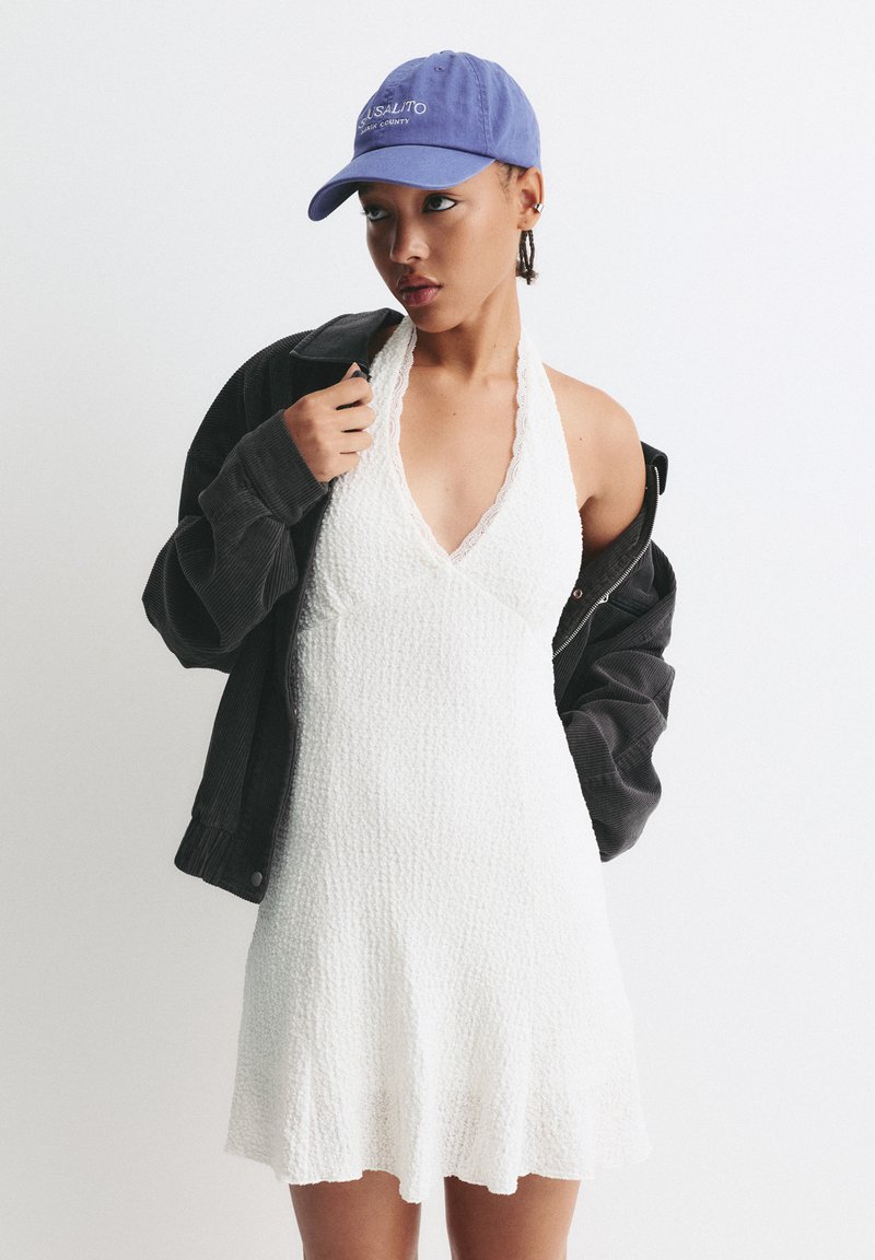 Robe halter blanche texturée avec dentelle, associée à une veste en velours côtelé noire et une casquette de baseball bleue ornée de texte brodé.