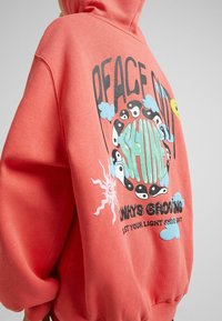 Sudadera color coral con un estampado gráfico en la parte posterior. Presenta texto estilizado, símbolos de yin-yang, nubes y acentos de sol en varios colores.