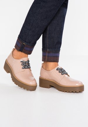 Lichtroze lakleren schoenen met een robuuste bruine plateauzool en zwarte veters met witte tekst. Enkellange jeans met kleurrijke omslag.