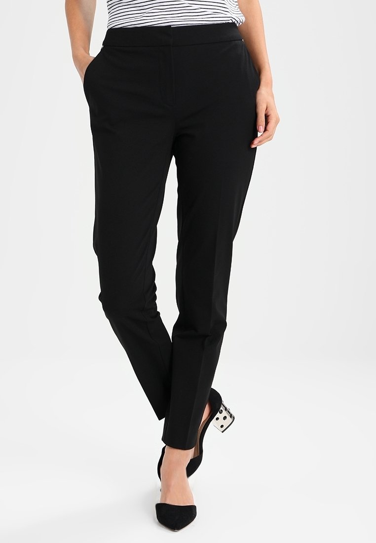 Femme portant un pantalon noir slim à la cheville avec poches latérales et des escarpins noirs à bout pointu avec talons blocs transparents ornés de pois noirs.