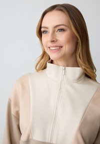 Beige en lichtbruine zip-up sweatshirt met hoge kraag en een zachte textuur; beschikt over contrasterende kleurvlakken en een traditionele ritssluiting.