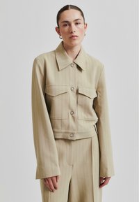 Beige pruhovaná zkrácená bunda s límcem, dvěma předními kapsami a knoflíkovým zapínáním. Tkanina má hladkou texturu a přiléhavý střih.