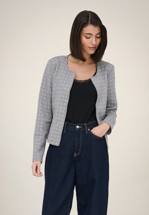 Femme aux cheveux courts et foncés portant une veste matelassée grise sur un haut noir orné de dentelle et un jean taille haute foncé, une main dans la poche.