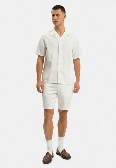 Hvid linned kortærmet skjorte og matchende shorts med knapfremspring. Båret med beige sokker og mørkebrune loafers.