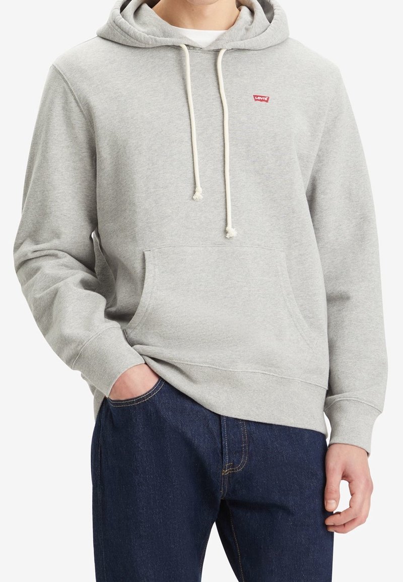 Adulte portant un sweat à capuche Levi's gris clair avec poche avant et cordons, associé à un jean bleu foncé, main dans la poche.