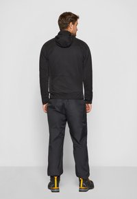 Giacca nera impermeabile con cappuccio, abbinata a pantaloni neri dal taglio ampio. I pantaloni hanno una finitura opaca elegante e stivaletti robusti.